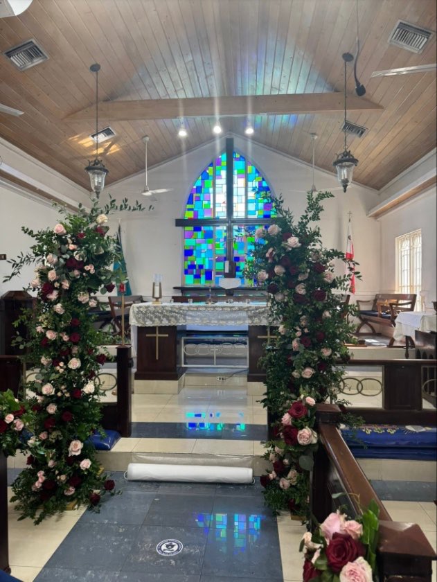 Tall rose and greenery arrangements frame a church aisle leading to a stained-glass window.